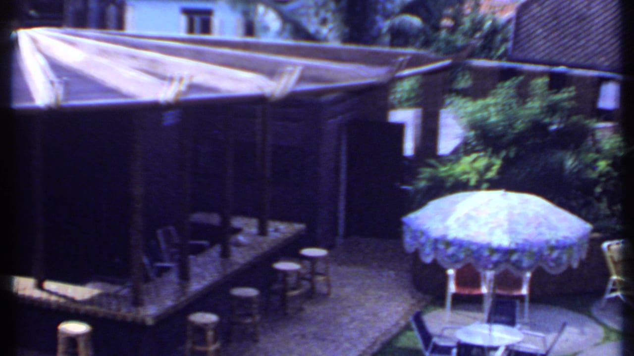 Sunlit courtyard with open blue umbrella, stools, lounge chairs, plants, and shaded bar area.