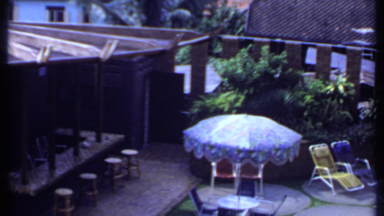 1969 India 8mm frame of a sunlit poolside-style courtyard with a blue umbrella, lounge chairs, stools, greenery, and brick buildings.