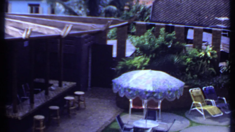 1969 India 8mm frame of a sunlit poolside-style courtyard with a blue umbrella, lounge chairs, stools, greenery, and brick buildings.
