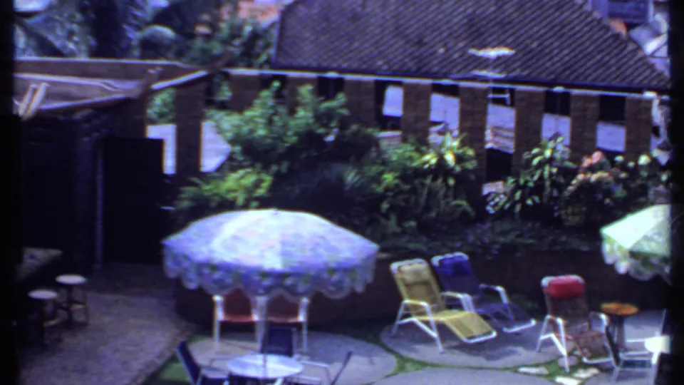 1969 India 8mm frame showing a blue umbrella shading chairs in a courtyard with lounge seating, small tables, greenery, brick wall, and tiled roof.