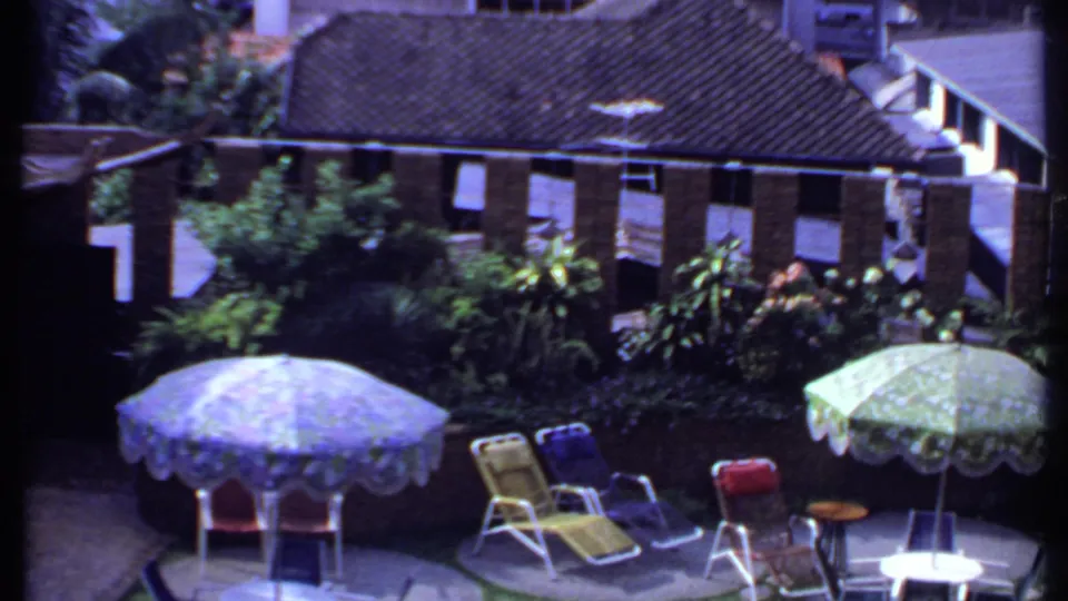 1969 India 8mm frame with blue and green umbrellas, lounge chairs, small round tables, dense plants, and brick buildings in bright sun.