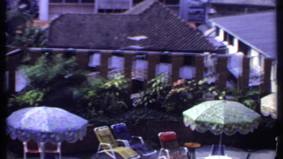 1969 India home-movie view of an outdoor patio with umbrellas, lounge chairs, dense shrubs, and adjacent buildings.