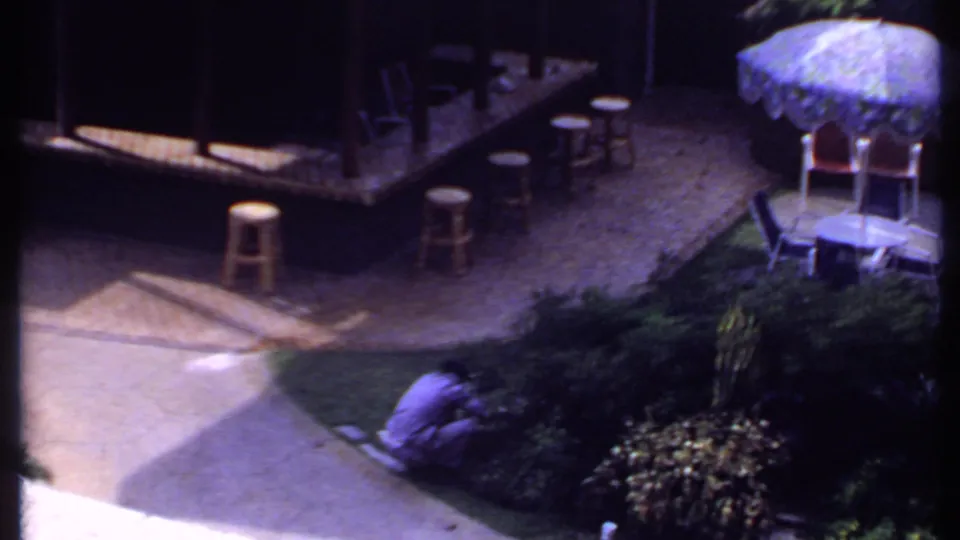 1969 India home-movie frame with a man tending shrubs near a curved walkway, stools, umbrella seating, and dense garden plants.