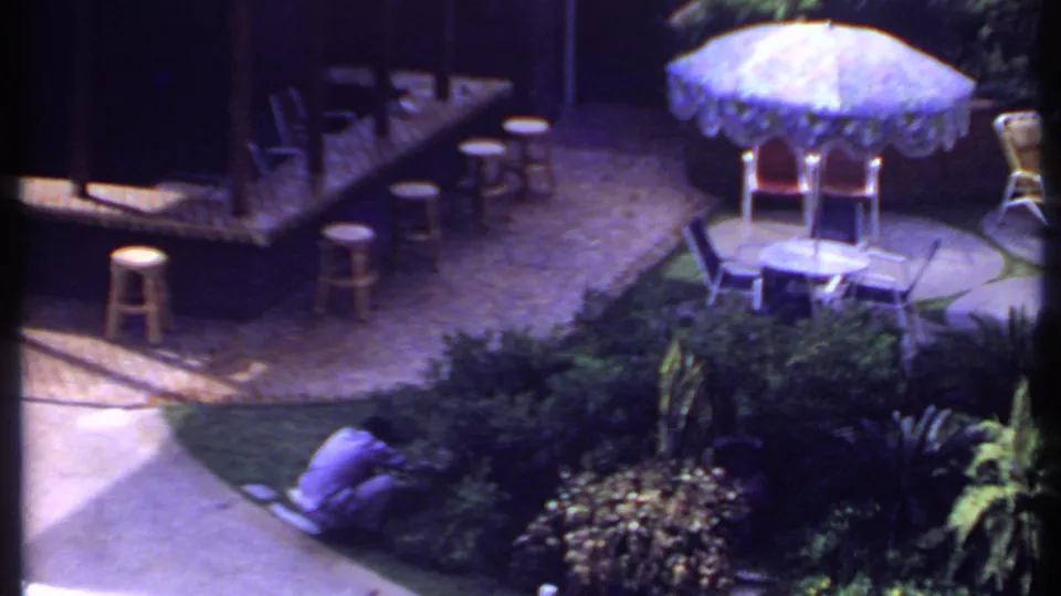 1969 India patio view showing a patterned umbrella, stools, chairs, dense shrubs, and a man crouched along the walkway.