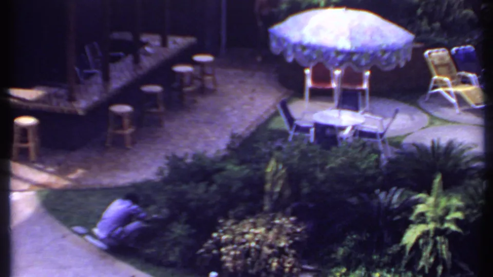 1969 India 8mm frame showing a tropical garden patio with stools, a fringed umbrella, lounge chairs, and a person crouched near foliage.