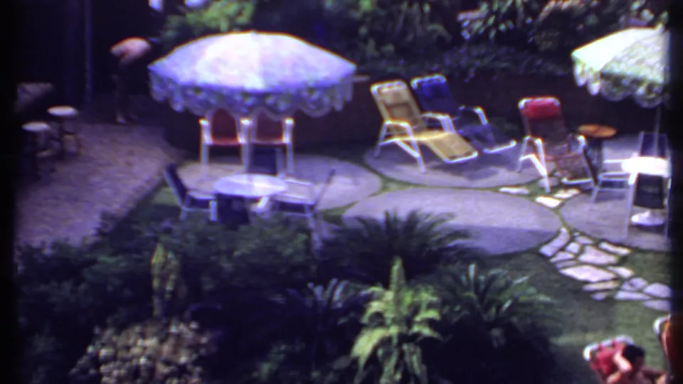1969 India 8mm frame showing the pan across a garden retreat with umbrella seating, lounge chairs, a stone path, and partially visible people.