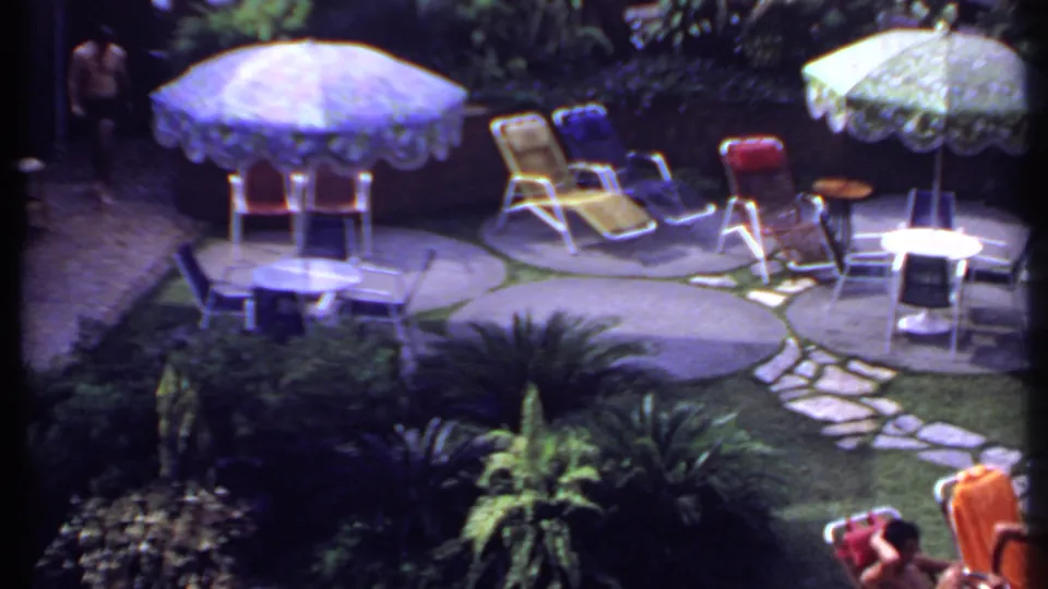 1969 India home-movie frame with umbrellas, colorful lounge chairs, garden planting, a stone path, and a partially visible seated person.