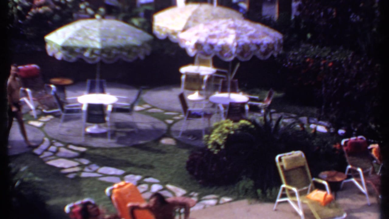 Sunlit patio with round tables, patterned umbrellas, chairs, and people at the edges.