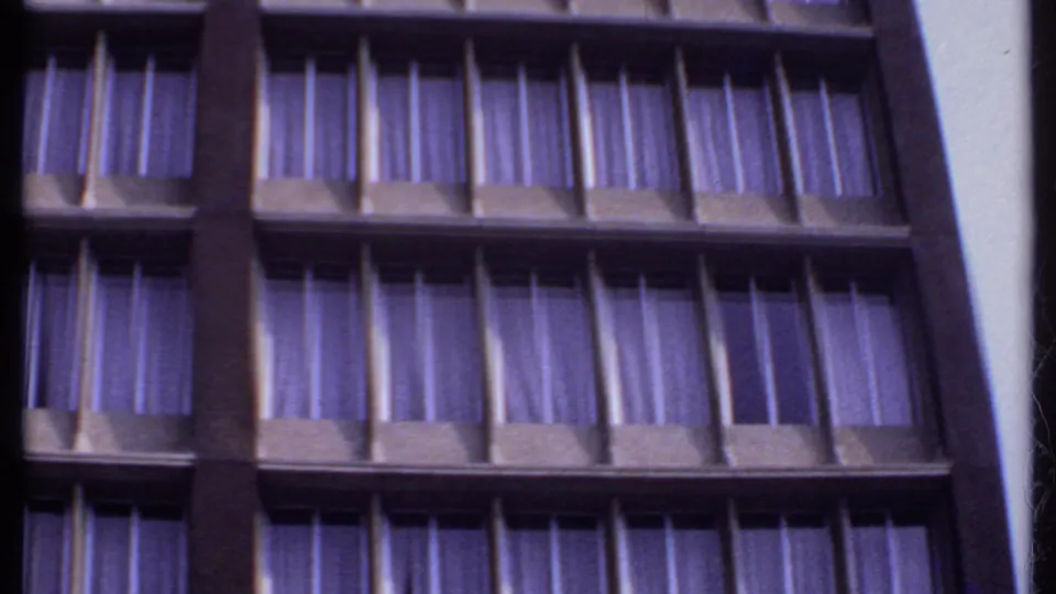 1969 India home-movie frame of a multi-story facade, emphasizing rectangular window bays and purple curtains.