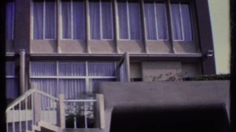 1969 India 8mm frame showing lower motel rooms with curtained windows, concrete balcony divisions, a small balcony rail, and a planter edge.