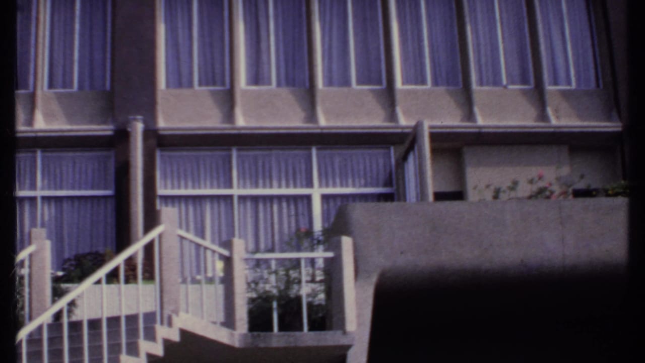Concrete building facade with curtained windows, balcony plants and white stair railings.