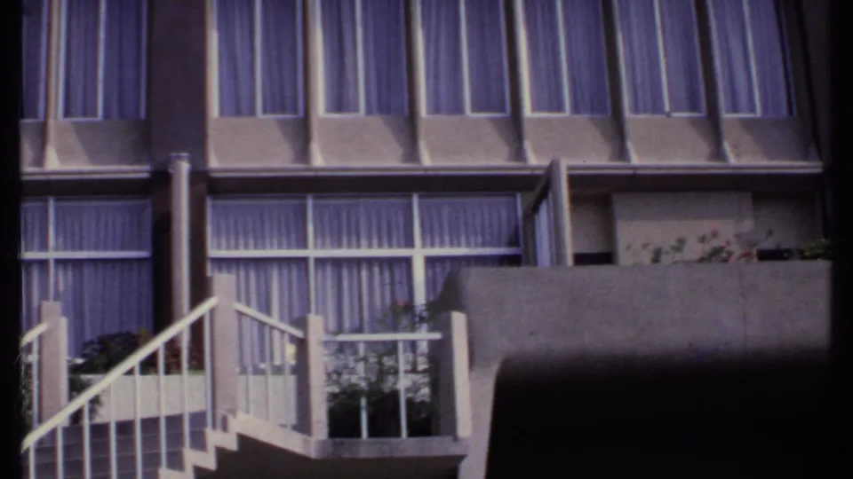 1969 India 8mm frame showing a modern concrete facade with purple curtains, balcony planters and white exterior stair railings.