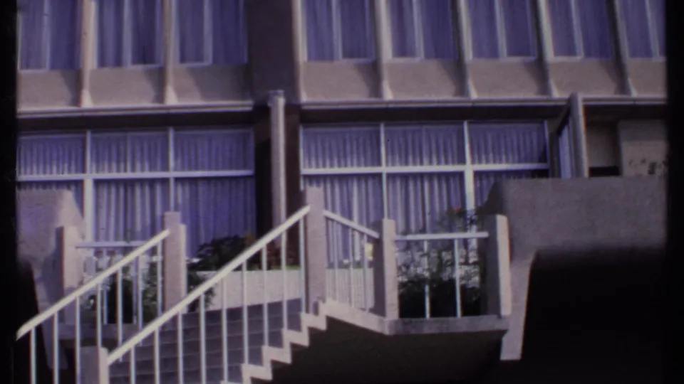 1969 India 8mm frame with a white-railed exterior staircase, balcony edge, planters and large windows behind curtains.