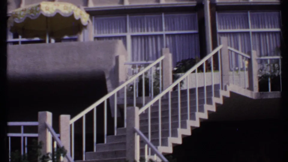 1969 India 8mm frame with a yellow umbrella or awning over a concrete balcony and white-railed stairs before curtained windows.