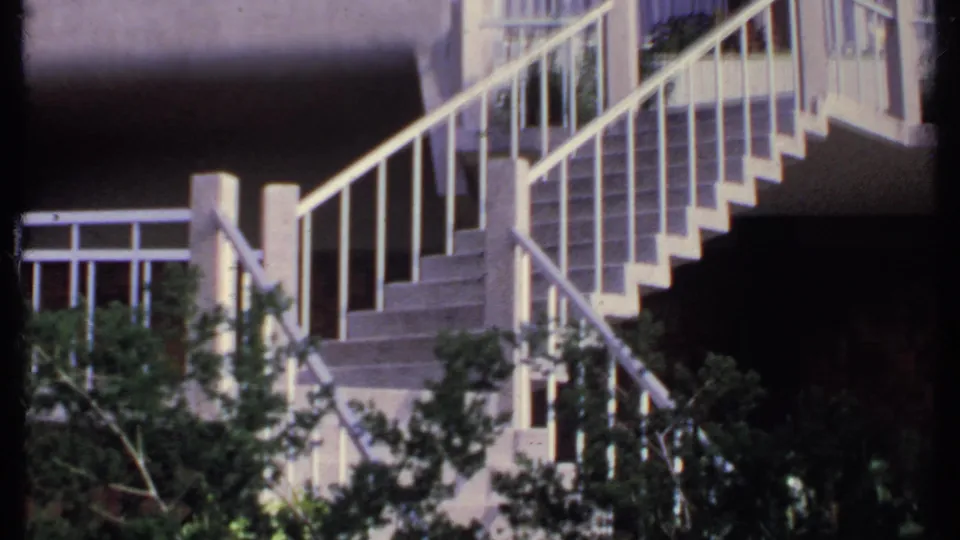 1969 India 8mm frame with a stair flight, pale railings, upper balcony windows, and leafy branches in the lower foreground.