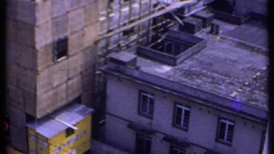 1969 India 8mm frame looking down across flat rooftops, a concrete building wall, small windows, and a yellow structure at lower left.