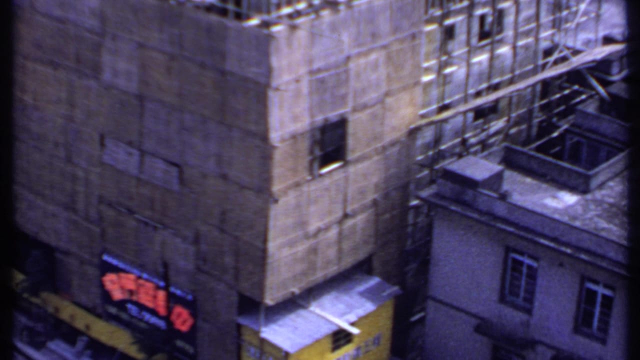 1969 India 8mm frame showing a concrete building under construction, scaffolding, nearby rooftops, and a red sign at street level.