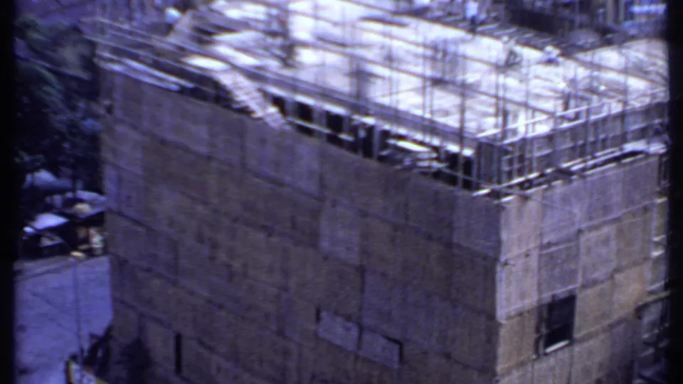 1969 India 8mm frame looking down on a rooftop work area, scaffold rails, concrete walls, and a narrow street beside the site.