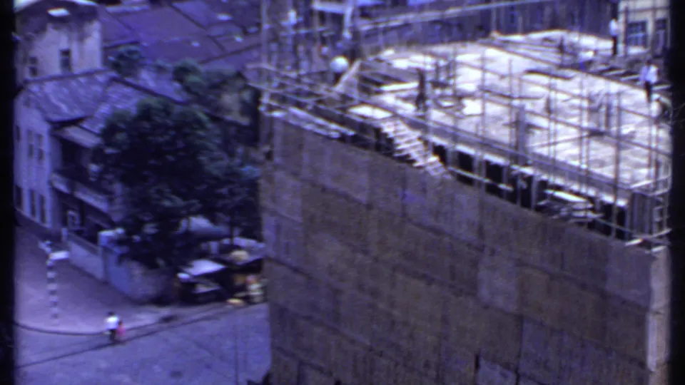1969 India 8mm frame showing a scaffolded rooftop construction site with nearby trees, low-rise buildings, and a small street scene below.