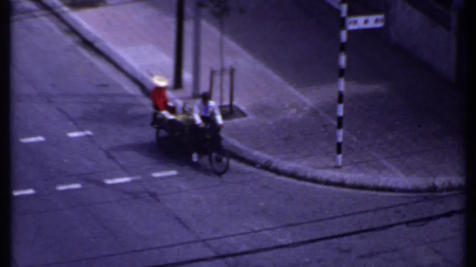 1969 India 8mm frame showing a red-shirted vendor on a rickshaw or cart beside a cyclist on a paved street corner.