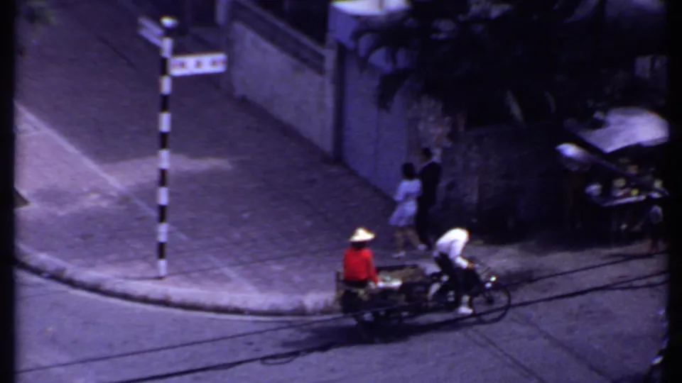1969 India home-movie frame showing a cart and cyclist in the road while a couple walks along the sidewalk near a wall.