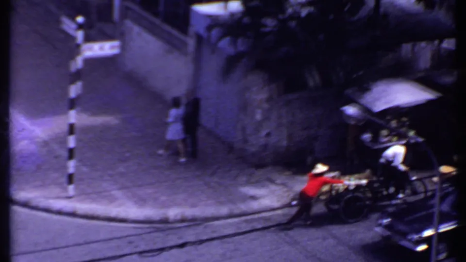1969 India street frame with the rickshaw or vendor cart at the lower right and pedestrians walking beside a shaded wall.