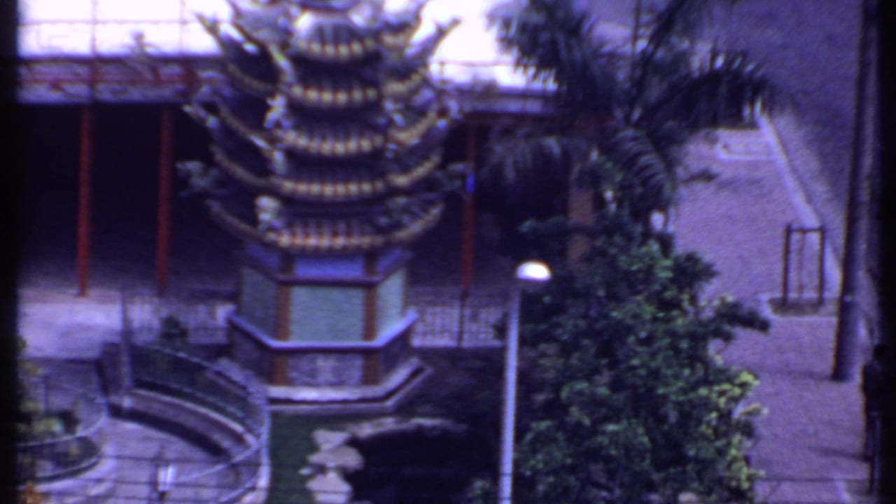 Pagoda-like tiered roof beside red columns, palm trees, courtyard paving, and a parked dark car.
