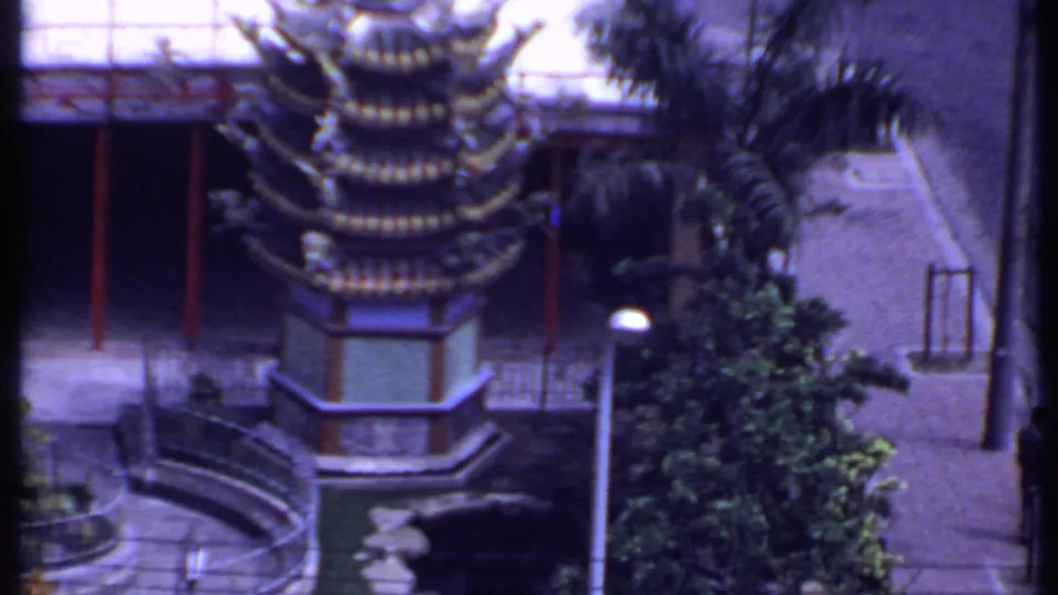 Elevated 1969 hotel-window view of a tiered pagoda-like roof, red columns, palm trees, courtyard paving, and a dark parked car.