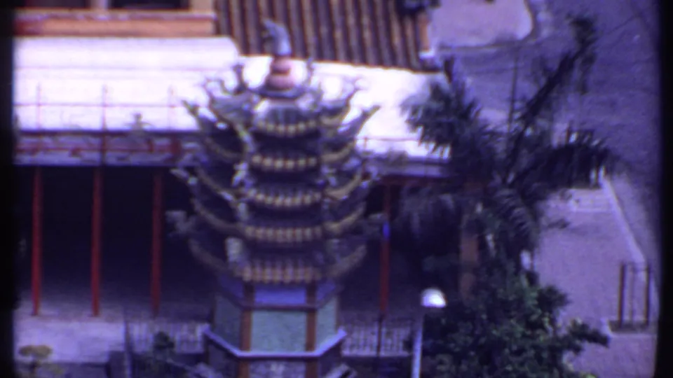 1969 8mm view looking down on a tiered Chinese roof form, red supports, palm fronds, railings, and a paved courtyard.