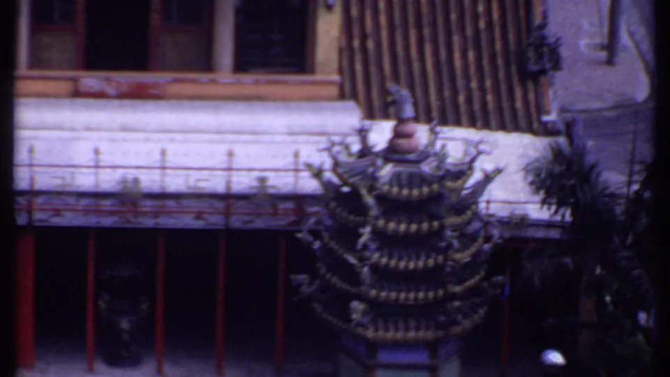 High-angle 1969 travel footage frame with a tiered roof, red-columned facade, tiled roofline, palm tree, and parked car below.