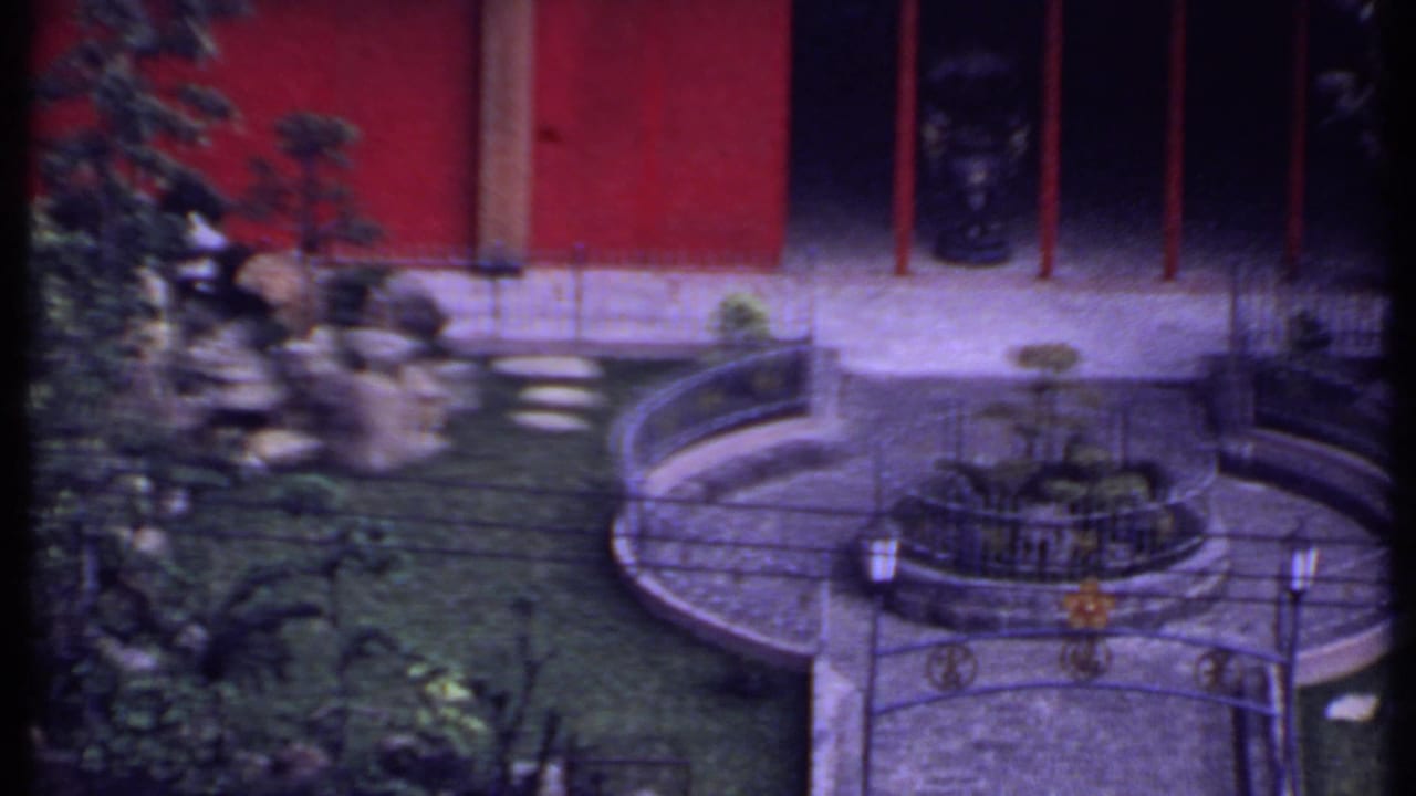 1969 India 8mm frame showing rocks, shrubs, stepping stones, and a curved courtyard railing against a red wall.