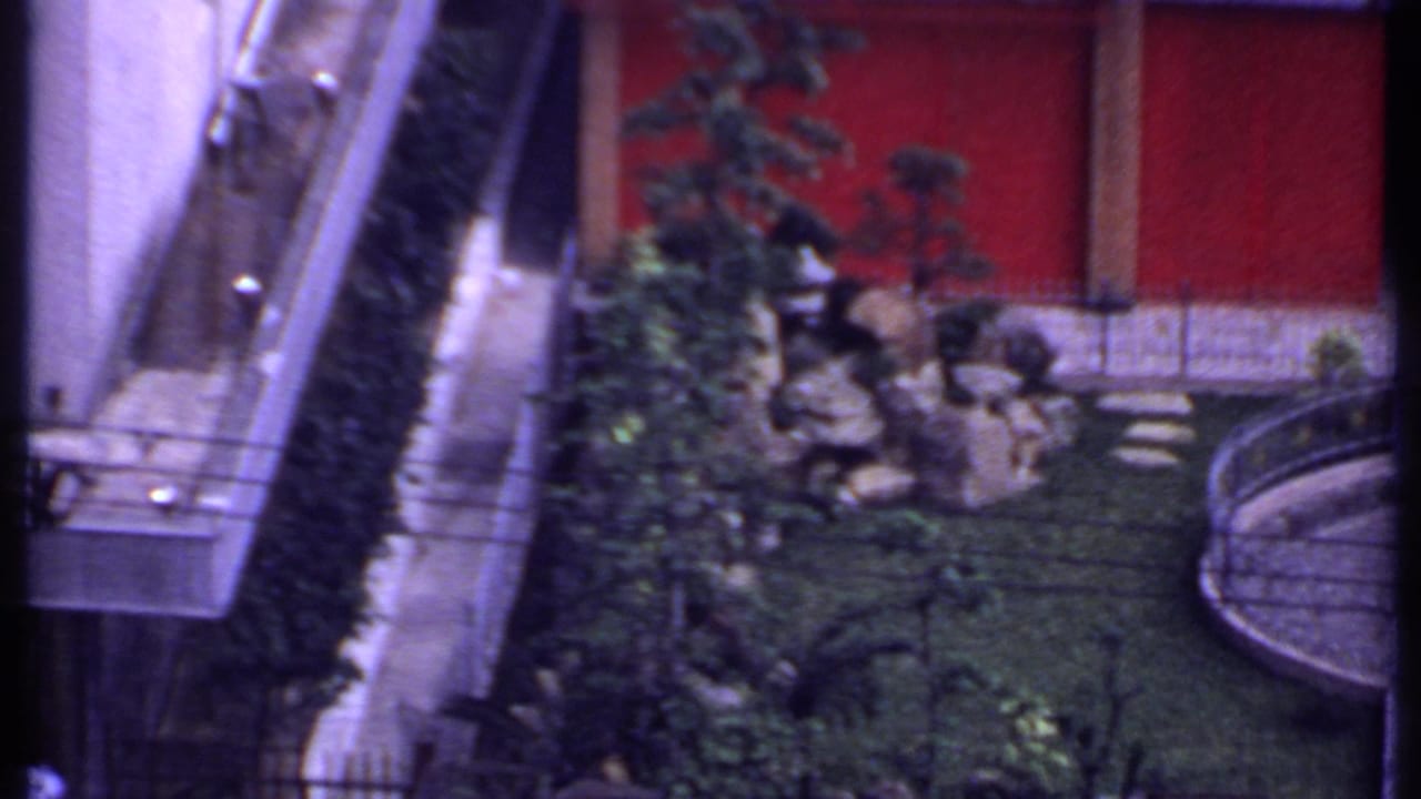 1969 India 8mm frame with a building edge, hedges, rocks, shrubs, red wall, and partial curved garden courtyard.