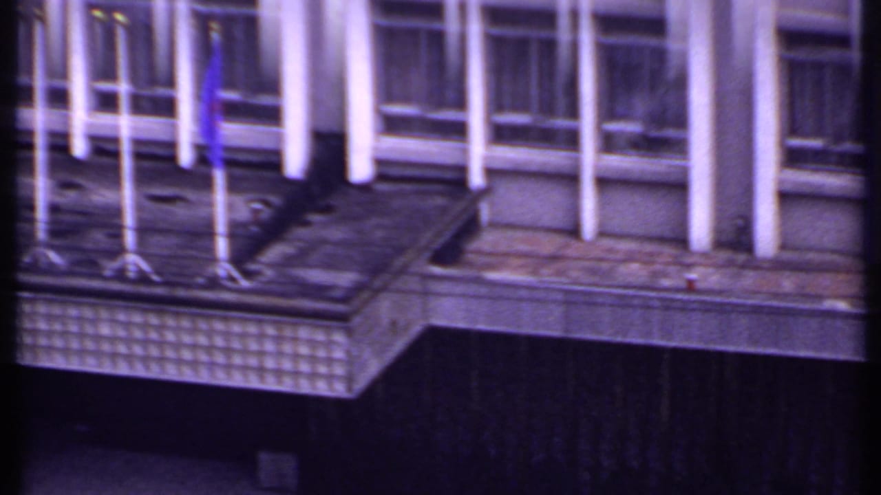 1969 India 8mm frame showing the building balcony with several flagpoles, a blue flag, windows, and white vertical columns.