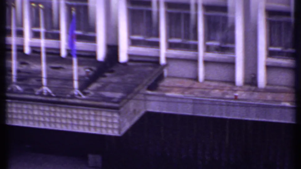 1969 India 8mm frame showing the building balcony with several flagpoles, a blue flag, windows, and white vertical columns.