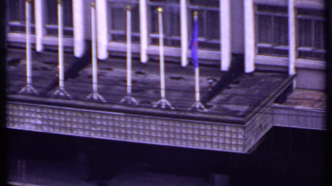 Rooftop edge with rows of empty flagpoles, one blue flag, large windows, and a tiled parapet.