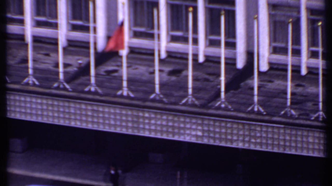 Building rooftop with many flagpoles, a red flag, large windows, and two distant figures below.