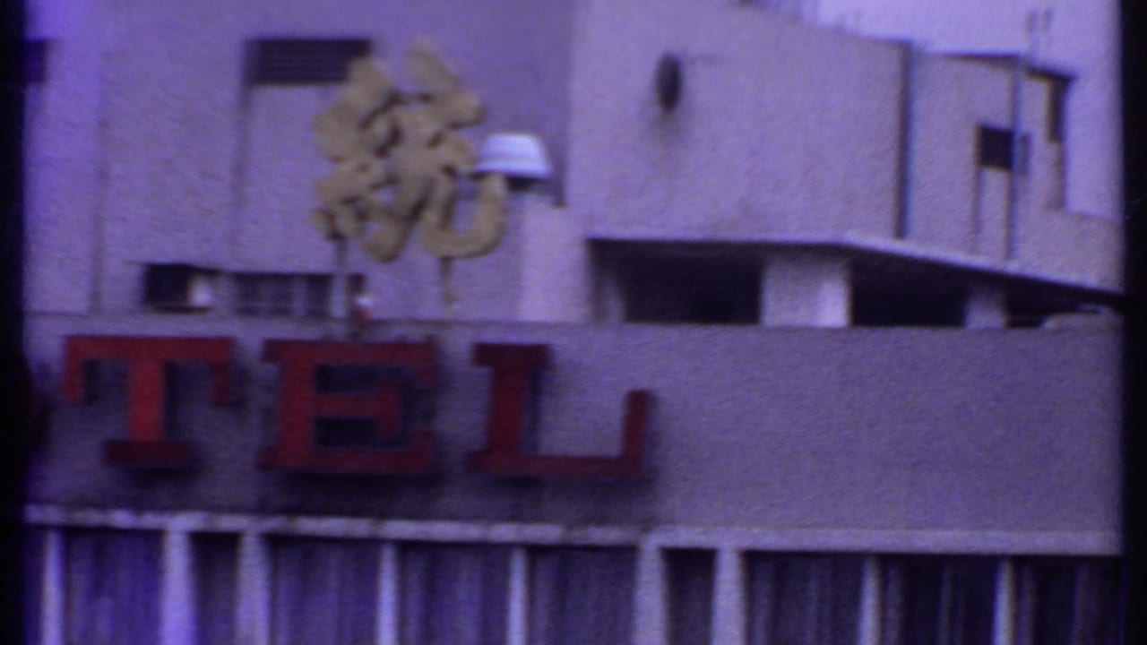 1969 India home-movie frame with partial TEL lettering, Chinese characters above, a pale hotel wall, and dark film edges.