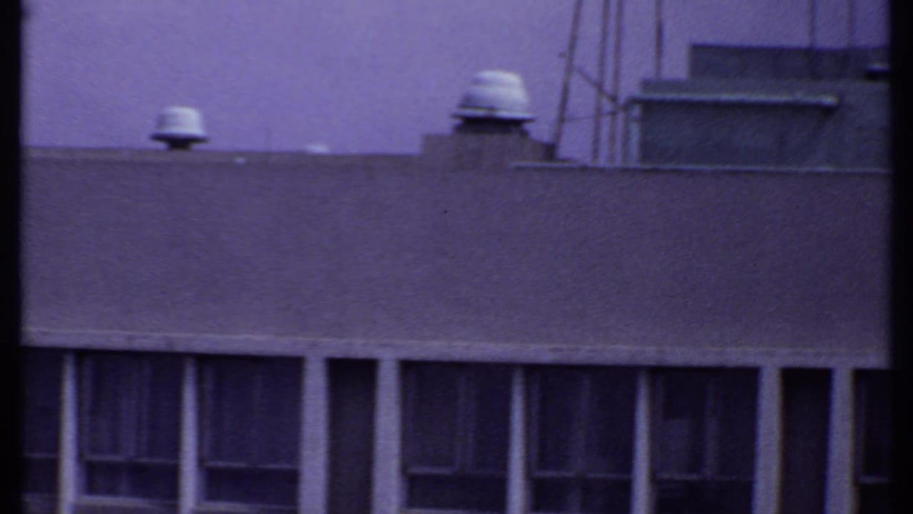 1969 India 8mm frame showing a flat industrial rooftop with vent caps, a boxy rooftop structure, and shadowed windows beneath.