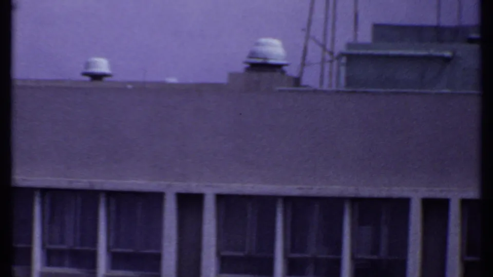 1969 India 8mm frame showing a flat industrial rooftop with vent caps, a boxy rooftop structure, and shadowed windows beneath.