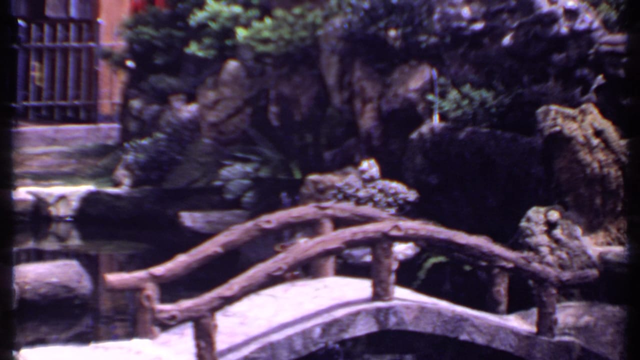 Arched wooden bridge over a pond with rocks, shrubs, and barred windows behind it.