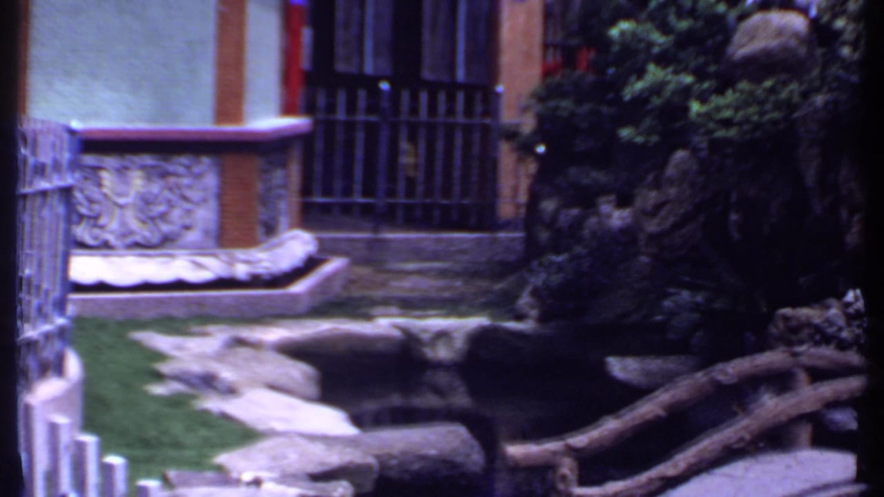 1969 India 8mm frame showing rock formations, a pond, bridge railing, shrubs, and barred windows in the garden setting.