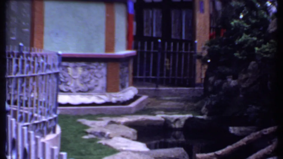 1969 India 8mm frame showing a courtyard with curved metal fencing, a carved stone basin, rocks, greenery, and a barred doorway.