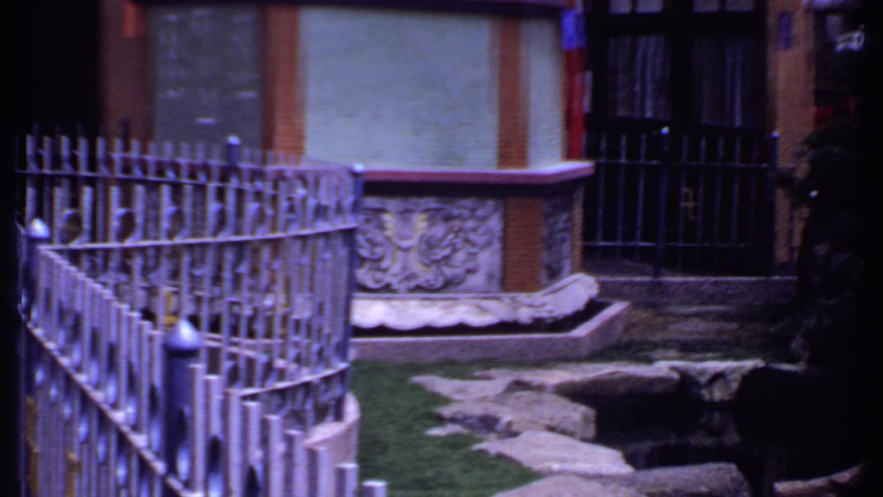 1969 India 8mm frame of a fenced courtyard area with a carved stone feature, rocks, greenery, and building facade.