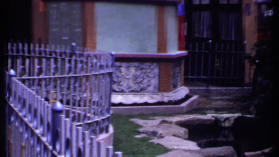 1969 India 8mm frame of a fenced courtyard area with a carved stone feature, rocks, greenery, and building facade.