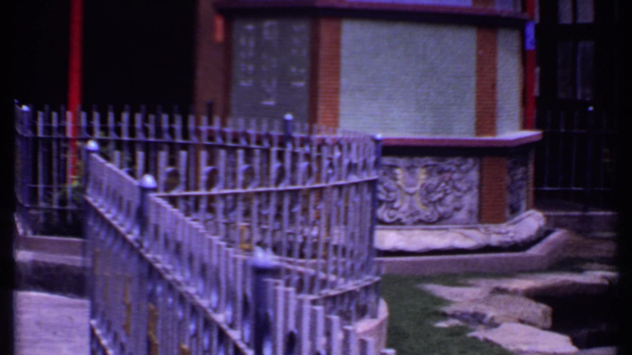 1969 India 8mm frame showing ornamental metal fencing, a barred doorway, carved wall detail, rocks, and a courtyard edge.