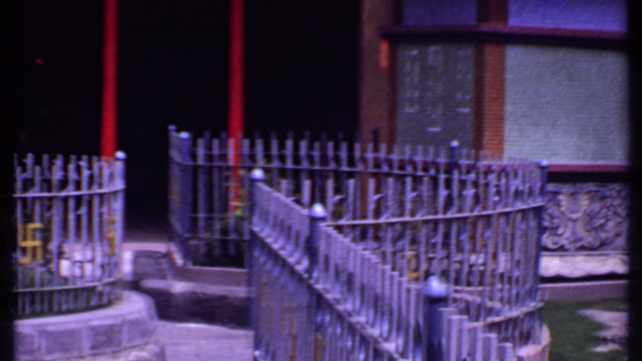 1969 India 8mm frame of a courtyard corner with curved metal fence, red post, shaded doorway, painted wall, and carved stonework.