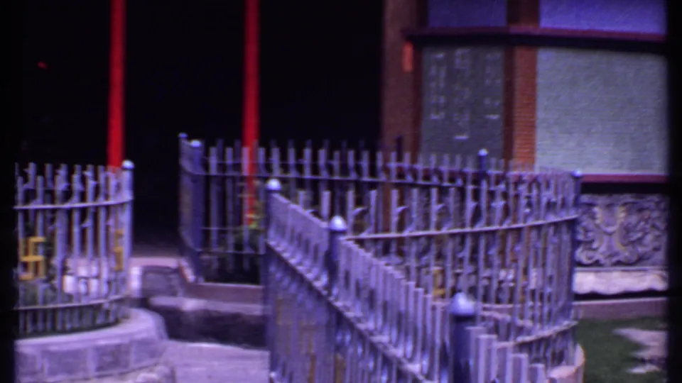 1969 India 8mm frame of a courtyard corner with curved metal fence, red post, shaded doorway, painted wall, and carved stonework.