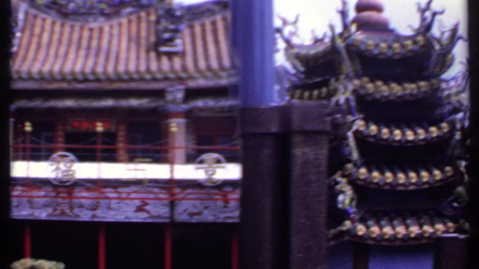 1969 India frame of a Chinese-style temple exterior, showing a tiled roof, red balcony railings, circular ornaments, and dark foreground columns.