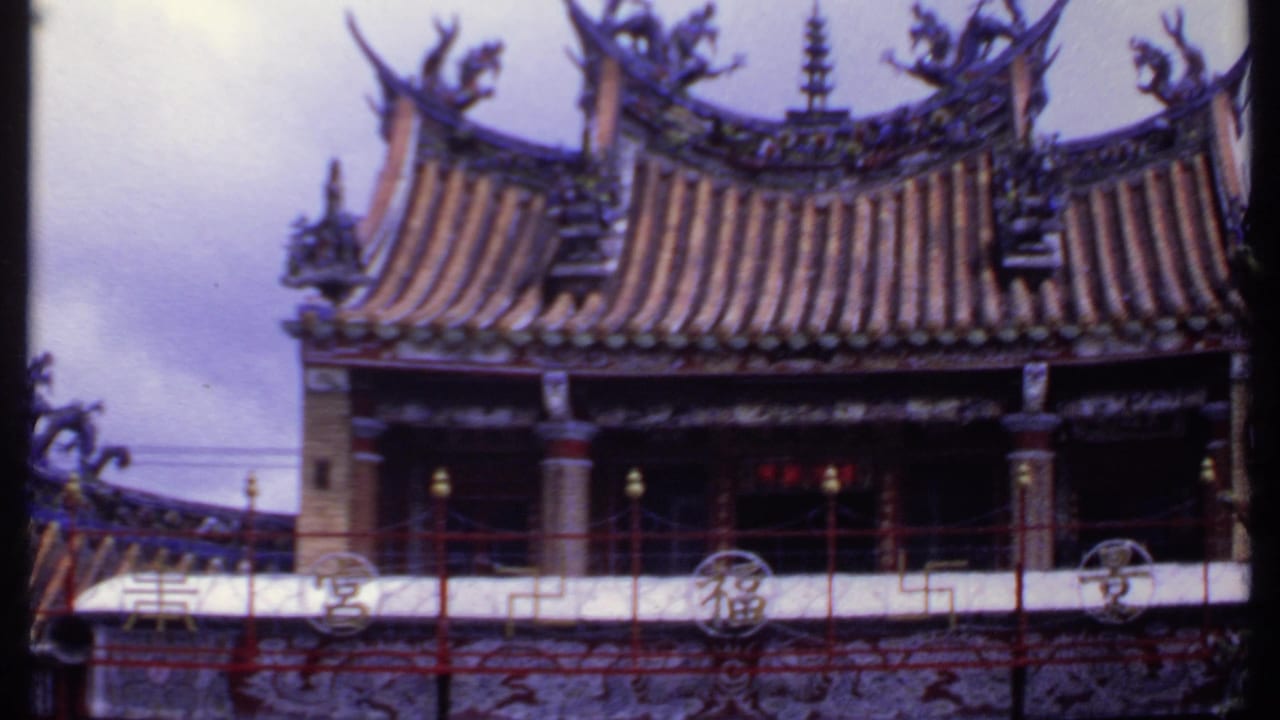1969 India frame showing a wider temple facade with upturned eaves, layered tile roof, red balcony railings, and cloudy sky behind it.