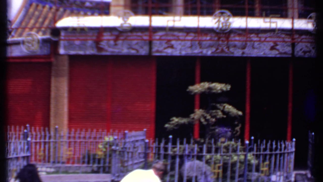 1969 India-listed 8mm view of a temple facade with red shutters, metal courtyard fence, small shaped tree, and partially visible visitors near the frame edge.